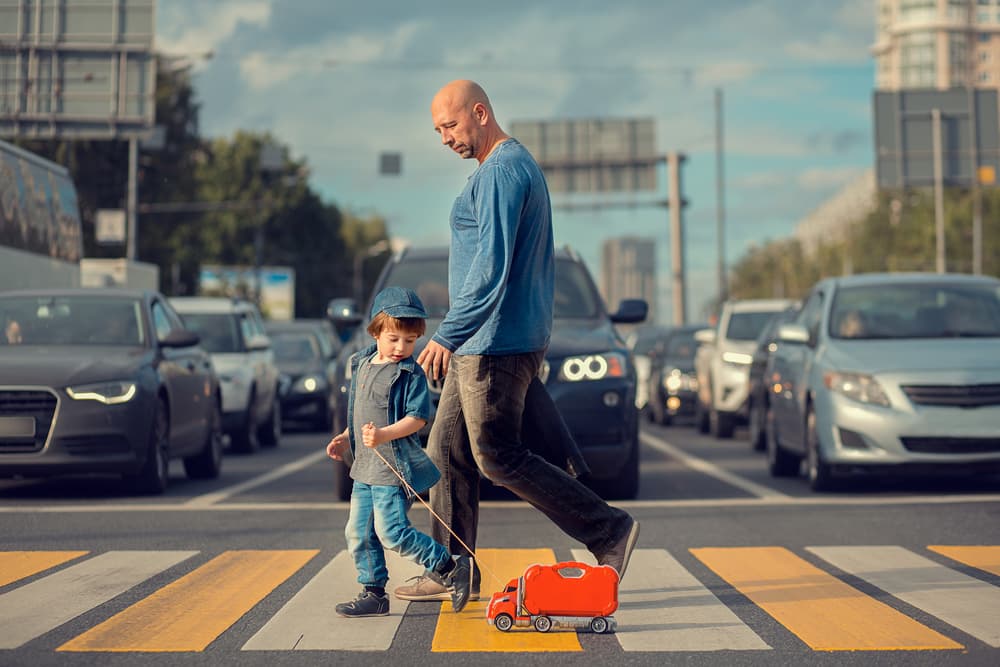 Young boy crossing street with father and red toy track, cars waiting at crosswalk. Selective focus.